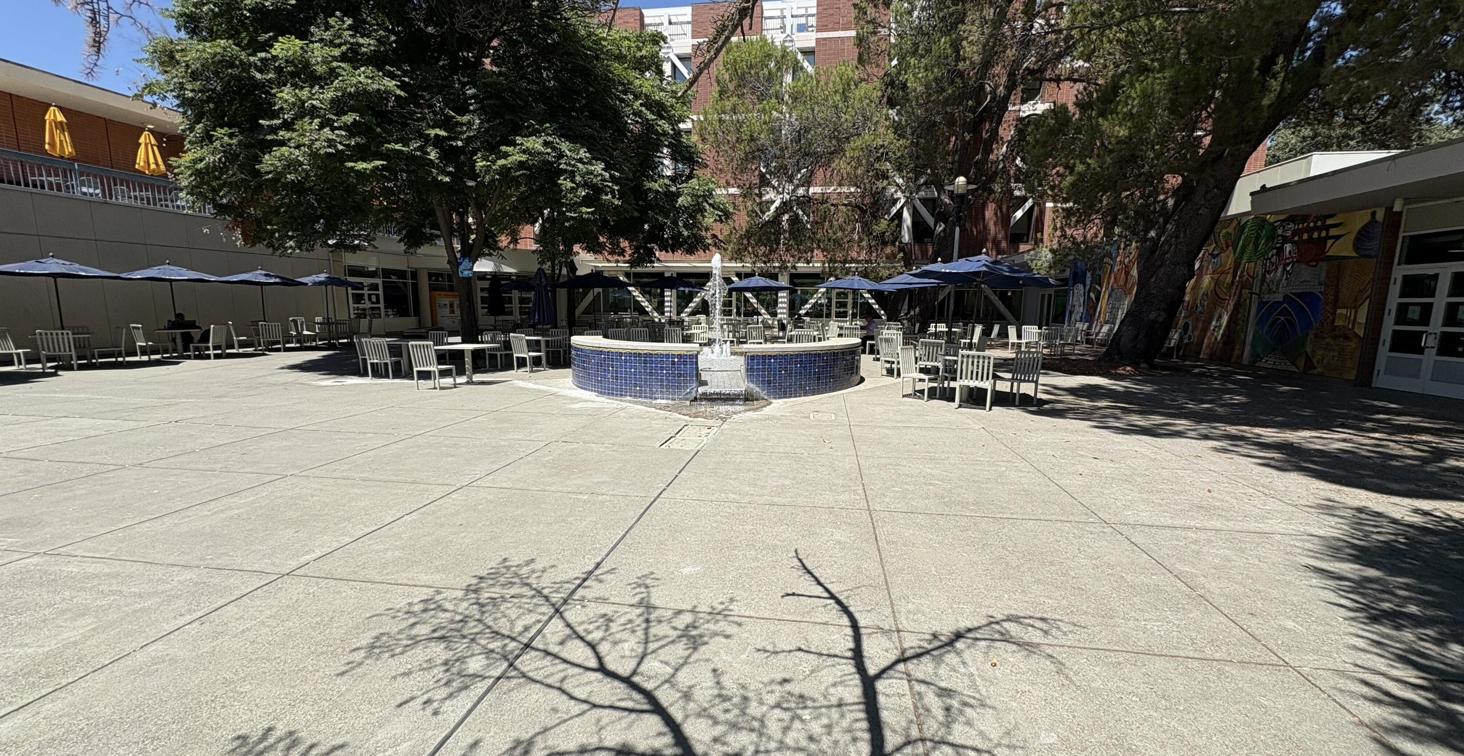A sunlit outdoor courtyard with a central blue-tiled fountain surrounded by white chairs and tables. Several trees provide shade, and large blue umbrellas are placed over some seating areas. In the background, there is a multi-story brick building partially obscured by tall trees. To the right, a colorful mural is visible on the wall of a building, and to the left, yellow umbrellas are seen on an upper patio area. The concrete ground is clean and spacious, with shadows of tree branches cast across it.